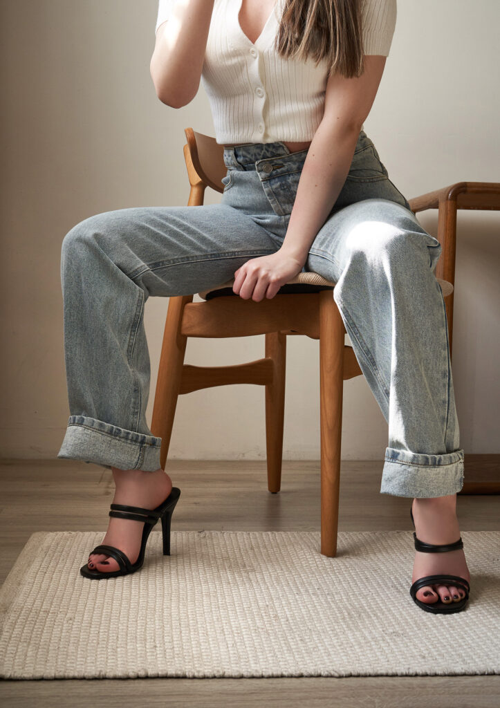 Gabriela Inzunza for Avice. A model sits on a wooden chair wearing a white button-down top and blue jeans paired with black strappy heels. The setting is minimalistic with a beige rug on a light wooden floor.